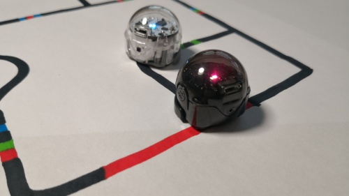 Die Szene zeigt zwei kleine, kugelförmige Roboter, die auf einem weißen Blatt positioniert sind. Der Roboter links ist transparent und der rechts ist tiefschwarz. Beide Roboter stehen auf einem weißen Hintergrund, auf dem schwarze Linien, unterbrochen von