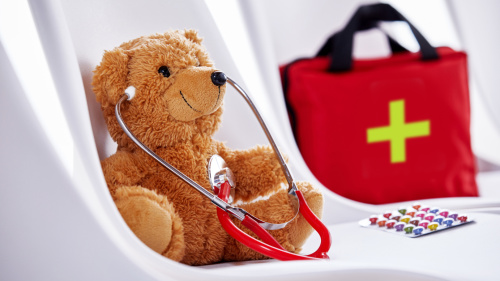 Das Bild zeigt einen braunen Teddybären, der auf einem weißen Stuhl sitzt und ein rotes Stethoskop um den Hals trägt. Im Hintergrund steht eine rote Erste-Hilfe-Tasche mit einem grünen Kreuz. Auf dem Stuhl liegt neben dem Bären eine Packung bunter Pillen.