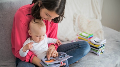 Das Bild zeigt eine junge Frau, die auf einem Sofa sitzt und ihrem Baby ein kleines Stoffbuch vorliest. Das Baby sitzt auf ihrem Schoß und scheint aufmerksam zuzuhören. Im Hintergrund liegen weitere Stoffbücher in einem Stapel.