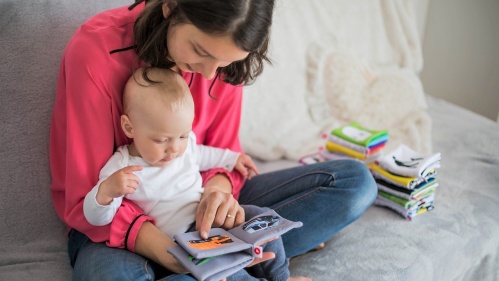 Das Bild zeigt eine junge Frau, die auf einem Sofa sitzt und ihrem Baby ein kleines Stoffbuch vorliest. Das Baby sitzt auf ihrem Schoß und scheint aufmerksam zuzuhören. Im Hintergrund liegen weitere Stoffbücher in einem Stapel.