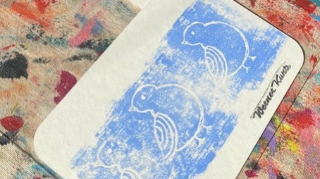 Das Bild zeigt zwei mit Tiefdruck hergestellten Postkarten auf einer bunten Malunterlage. Die obere Postkarte zeigt einen Druck einer Blume, dabei sind die Umrisse der Blume ausgespart und der Bereich darum mit roter Farbe ausgefüllt. Das untere Bild zeig