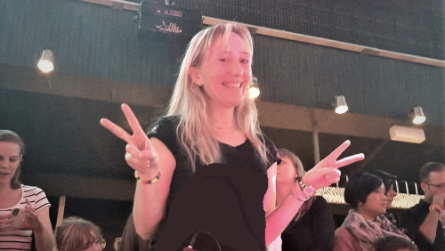 Das Bild zeigt eine junge, lächelnde Frau mit langen, blonden Haaren, die in einer Halle steht und das Victoryzeichen macht, indem sie bei angewinkelten Armen Zeige- und Mittelfinger beider Hände zu einem V formt. Die Frau trägt bequeme Tanzbekleidung: ei