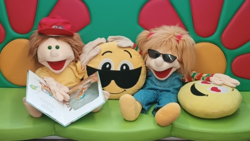 Das Bild zeigt eine gemütliche Leseecke in einer mobilen Bibliothek, dem BI-BUS der Stadtbibliothek Saarbrücken. Im Mittelpunkt sitzen zwei Handpuppen auf einem grünen Sitz: Lucien, eine Puppe mit roter Mütze, die ein Buch liest und Maja, eine Puppe mit S