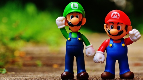 Das Bild zeigt zwei Plastikfiguren von Super Mario und Luigi. Sie stehen auf einem braunen Untergrund vor einem verschwommenen Hintergrund aus grünem Gras. Beide Figuren heben eine Hand zum Gruß oder in Siegerpose.