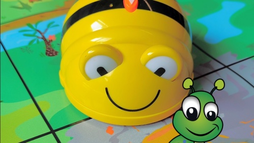Das Bild zeigt einen kleinen Roboter in Gestalt einer Biene. Er ist gelb mit schwarzen Streifen, großen Augen und einem lächelnden Mund. Davor sieht man eine gezeichnete Raupe. Beide Figuren befinden sich auf einer Matte, auf der eine Landschaft abgebilde