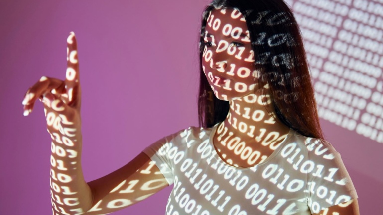 Vor einem violetten Hintergrund steht eine Frau, auf deren Gesicht, Körper und erhobene Hand ein binärer Code projiziert wird.