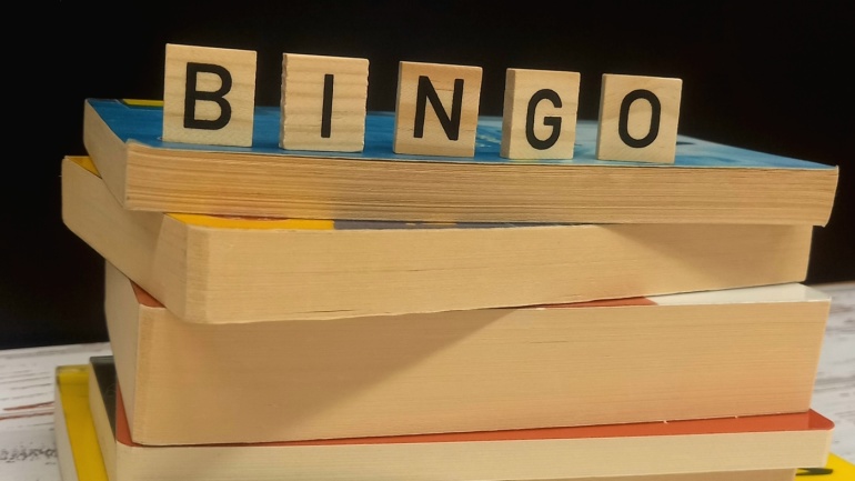 Vier gestapelte Bücher mit hölzernen Buchstabenkacheln, auf denen „Bingo“ steht, vor einem dunklen Hintergrund.