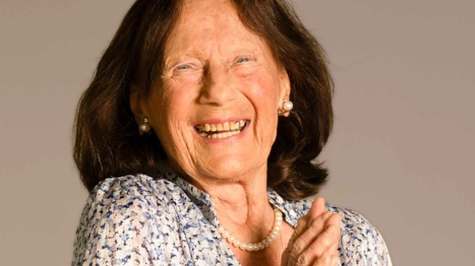 Eine ältere Frau mit langen braunen Haaren lächelt und faltet die Hände. Sie trägt eine geblümte Bluse, eine Perlenkette und Perlenohrringe vor einem einfarbigen Hintergrund. Eine ältere Frau mit langen braunen Haaren lächelt und faltet die Hände. Sie trägt eine geblümte Bluse, eine Perlenkette und Perlenohrringe vor einem einfarbigen Hintergrund.