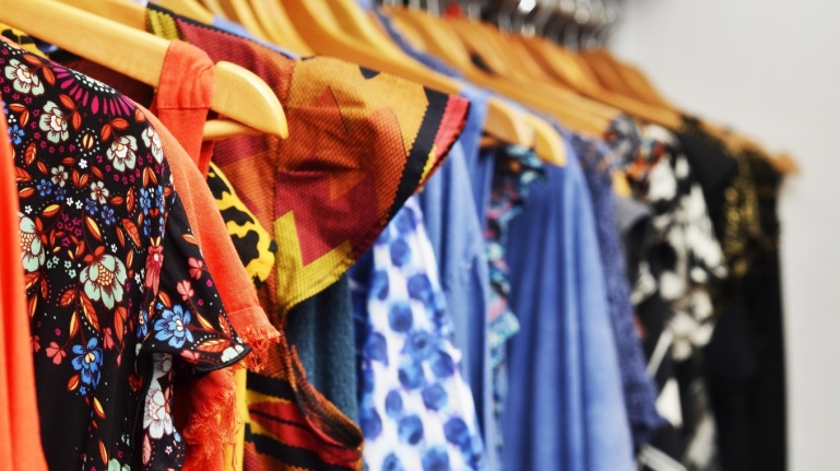 Nahaufnahme von farbenfrohen gemusterten und einfarbigen Hemden und Kleidern, die auf Kleiderbügeln in einem Bekleidungsgeschäft hängen.