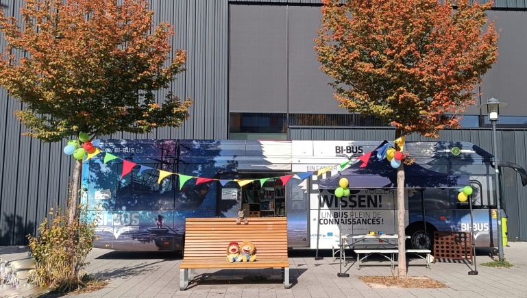 Der Bücherbus auf dem Gelände des Eurobahnhofs, im Vordergrund zwei Bäume mit Herbstlaub Der Bücherbus auf dem Gelände des Eurobahnhofs, im Vordergrund zwei Bäume mit Herbstlaub