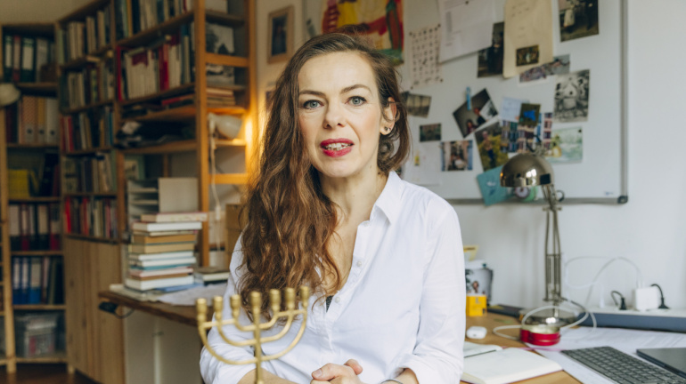 Eine Frau mit langen Haaren und einem weißen Hemd sitzt in einem unaufgeräumten Heimbüro, hinter ihr an der Wand hängen Bücher, Papiere und Fotos. Eine Frau mit langen Haaren und einem weißen Hemd sitzt in einem unaufgeräumten Heimbüro, hinter ihr an der Wand hängen Bücher, Papiere und Fotos.